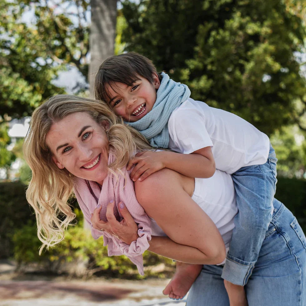 Frau mit Kind im Arm, beide tragen Musselin-Halstücher in Rosa und Hellblau – fröhlicher Moment im Freien mit leichten Bio-Tüchern von Hutch&putch