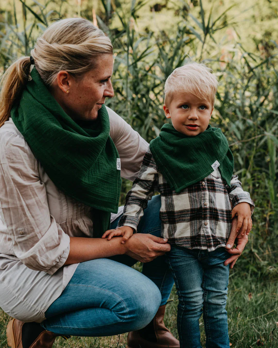 Eine Frau und ein kleiner Junge tragen beide grüne Musselin-Halstücher und stehen im Grünen; sie schauen sich liebevoll an.